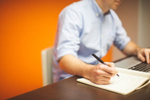 a caucasian man writing on a notepad while in front of a laptop