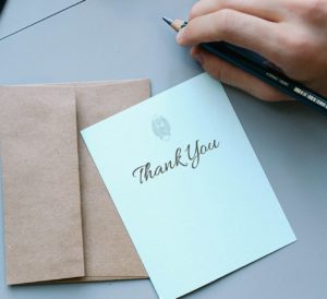 caucasian hand with a pen in it next to a white card that says thank you