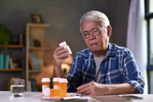 older asian man looking at a pill bottle in his hand with other bottles sitting on the table in front of him