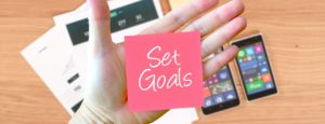 set goals written on a post it note wit cell phones and paper in the background