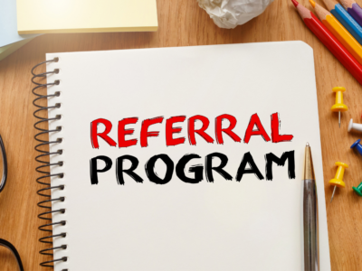 A notebook with the phrase "Referral Program" written in bold red and black letters, surrounded by colorful office supplies on a wooden desk.