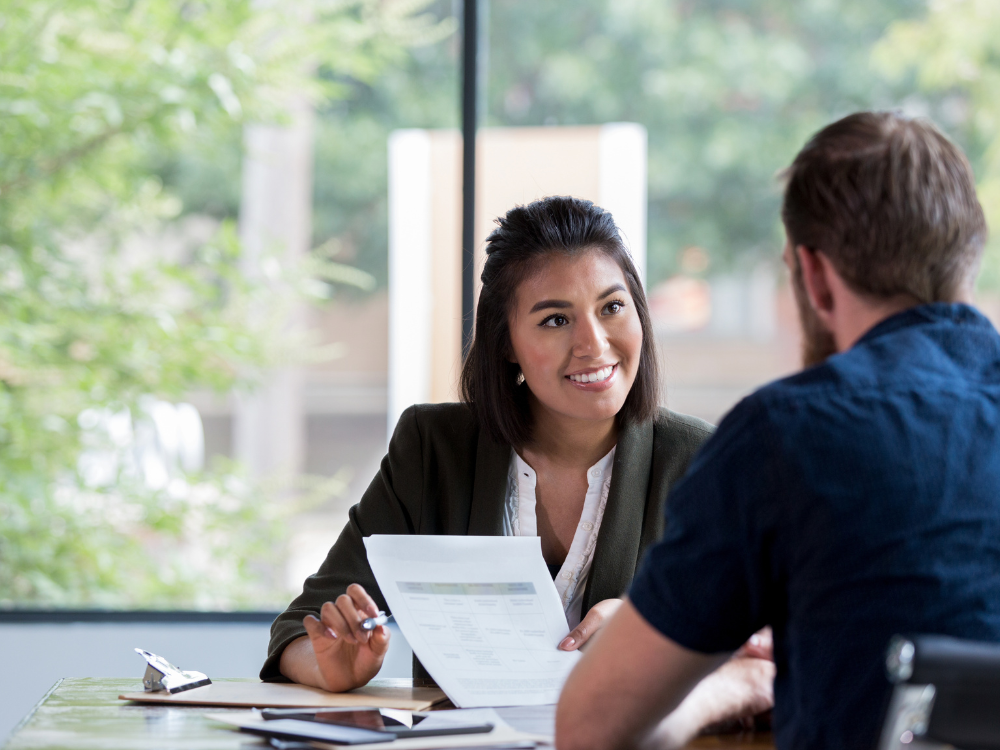 A female insurance agent smiling and holding a document while meeting with a client, symbolizing trust and personal connection in insurance sales.