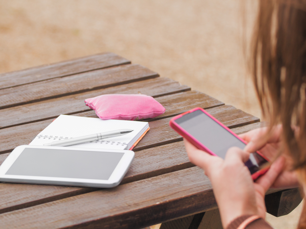 A person using a pink smartphone outdoors at a wooden table, with a planner, pen, and tablet beside them.